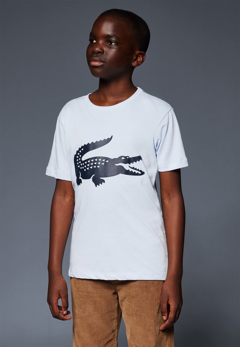 T-shirt en coton bleu clair avec un grand graphique de crocodile noir. Manches courtes et design à col ras du cou, associé à un pantalon en velours côtelé marron.