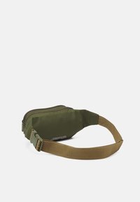 Olivgrön tygmidjeväska med en dragkedja och ett brunt justerbart band. Har en minimalistisk design och logotypdetalj på sidan.