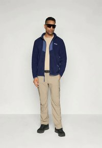 Marine-fleecejakke med lyseblå detaljer, beige skjorte, beige cargo-bukser og svarte støvler. Solbriller på, hender ved siden.