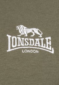 Olive zelená tkanina so soft textúrou obsahuje biele vyšívané logo leva a "Lonsdale London" v tučnom, zakrivenom písmene.