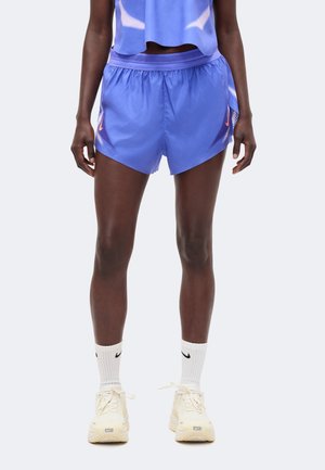 Person trägt blaue Sportshorts, passendes ärmelloses Oberteil, weiße Nike-Socken und cremefarbene Nike-Laufschuhe und steht vor einem einfarbigen Hintergrund.