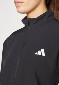 Sort atletisk jakke med høj krave, frontlynlås og hvidt logo. Glat tekstur og tætsiddende design, velegnet til aktivt tøj.