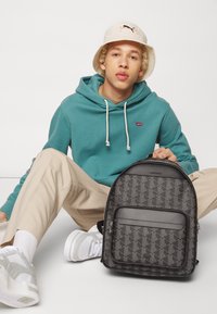 Rucsac negru cu un model geometric repetitiv, buzunar frontal cu fermoar și material sintetic, asortat cu un hanorac teal și pantaloni bej.