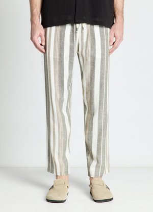 Homme portant un pantalon beige, gris et blanc à rayures verticales et des chaussures beige à enfiler, debout les mains relâchées le long du corps.