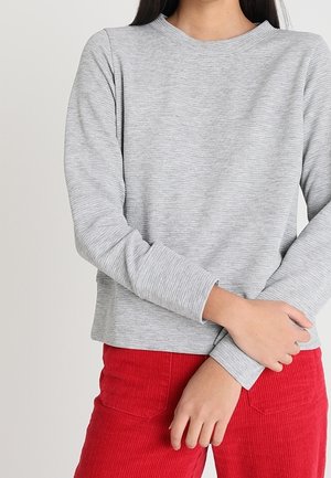 Osoba ubrana w jasnoszary, prążkowany sweter z długim rękawem i jaskrawo czerwone spodnie sztruksowe, z rękami skrzyżowanymi na talii.