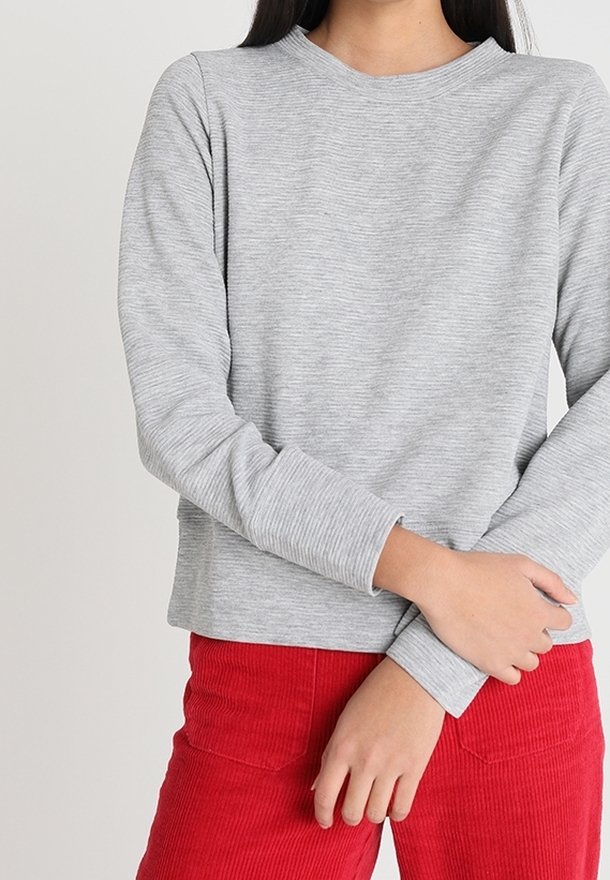Personne portant un pull côtelé gris clair à manches longues et un pantalon en velours côtelé rouge vif, bras croisés à la taille.