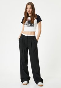 Korte, cropped wit-zwarte graphic t-shirt met korte mouwen; zwarte high-waisted geplooide broek; witte tailleband; model draagt witte sneakers.