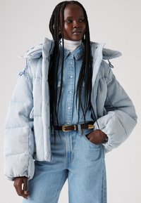 Light blue puffer jacket with large quilted sections, button and zip closure, worn over a matching denim shirt and high-waisted jeans.