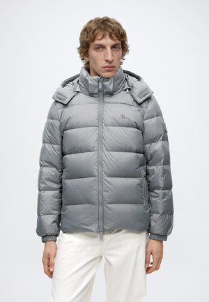 Jeune homme portant une doudoune gris clair avec col montant et capuche, associée à un pantalon blanc, debout devant un fond uni.