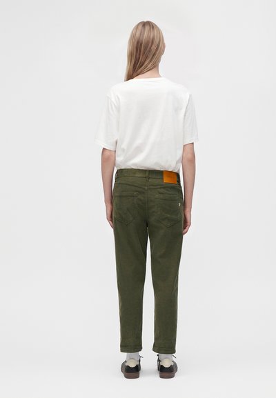 Personne portant un T-shirt blanc, un pantalon vert olive avec un patch marron, des chaussettes blanches et des chaussures noires, debout, vue de dos sur un fond uni.