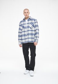 Grå og blå rutete flanellskjorte med knappestolpe foran, to brystlommer, parret med svarte bukser med hvite sømmer og hvite sneakers.