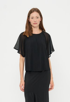 Femme aux cheveux bruns raides portant un haut noir plissé avec des manches courtes transparentes et une jupe noire, debout sur un fond blanc.