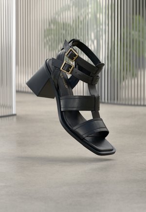 Schwarze Sandalen mit Absatz, die glatte Lederriemen und ein quadratisches Zehendesign aufweisen. Doppelte goldene Schnallen akzentuieren die Knöchelriemen.