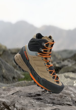 Középmagas, cserzett és fekete túrabakancs narancssárga cipőfűzővel és díszítésekkel, sziklás terepen, háttérben homályos hegyekkel.