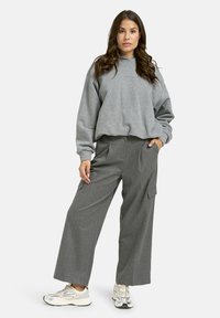 Eine Frau steht mit der Hand in der Tasche, trägt einen lockeren grauen Hoodie, weit geschnittene graue Hose und weiße Sneaker vor einem einfarbigen Hintergrund.