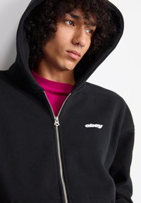 Black zip-up hoodie with a silver zipper and small white "obey" logo on the left side, worn over a fuchsia shirt. Soft fabric texture.