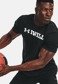 T-shirt sportivo nero con testo bianco "IWILL" e logo Under Armour; il indossatore tiene un pallone da basket rosso, mostrando la definizione muscolare del braccio.