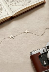 Silverhalsband med en rund hänge med ett globdesign och en fågelberlock, utställd på beige tyg tillsammans med en vintagekamera.