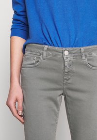 Szare dżinsy z denimu o dopasowanym kroju, wyposażone w zapięcie na guziki z przodu, boczne kieszenie oraz subtelny wzór faktury. Stylizowane z niebieskim swetrem.