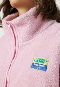 Roosa fliisjakki kõrge kaelusega, millel on tõmblukk ja silt, millel on kirjas "LA MAISON AMV SUPER VINTAGE" värvilises tekstis.