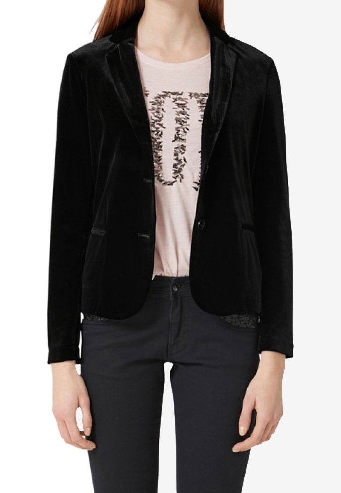 Femme portant un blazer en velours noir sur un T-shirt rose clair à imprimé noir, assorti à un pantalon noir, debout les bras détendus.