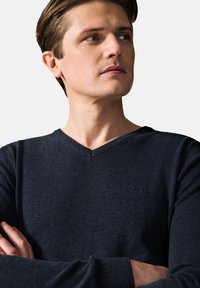 Granatowy sweter z dekoltem w serek, wykonany z teksturowanej tkaniny, z małym logo na lewej piersi i ściągaczami na rękawach. Prosty, nowoczesny design.