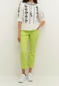 Femme portant un chemisier blanc avec une broderie florale noire et un pantalon court vert citron, debout avec une main dans la poche et des baskets blanches.