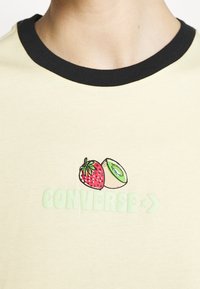 Jasnożółta koszulka z czarnym kołnierzykiem. Zawiera grafikę truskawki i kiwi, z napisem "CONVERSE" na zielono pod wzorem.