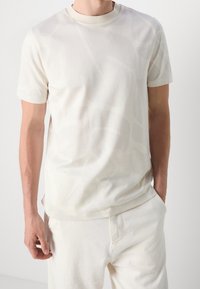 Manlig torso klädd i en kortärmad vit t-shirt med subtila abstrakta mönster och vita byxor, med en hand i fickan.