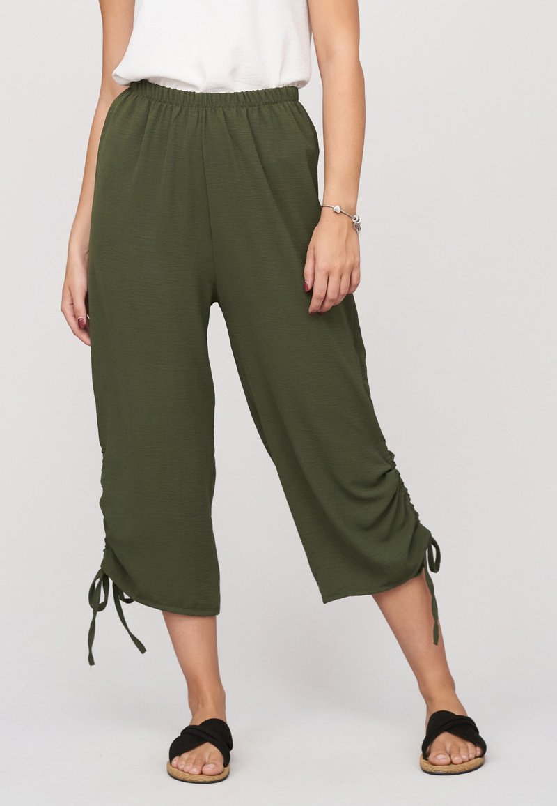 Femme portant un pantalon ample court vert olive avec cordons, un haut blanc et des sandales noires à enfiler avec des brides torsadées.