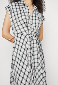 Šaty s knoflíky z lehké tkaniny s černo-bílým kostkovaným vzorem. Límeček, krátké rukávy a detail s převázáním v pase.