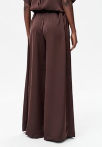 Bruine broek met wijde pijpen van gladde stof, met een losse pasvorm en een elastische tailleband, weergegeven van achteren.