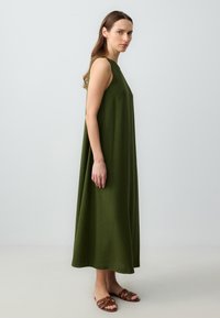 Bezrękawna, do kolan sukienka w głębokiej zieleni, wykonana z teksturowanej tkaniny. Swobodny krój z luźnym dopasowaniem, w połączeniu z brązowymi klapkami.