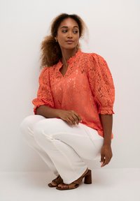 Coral lace blouse with a ruffled neckline and puffed sleeves, paired with white fitted pants and brown block-heeled sandals.