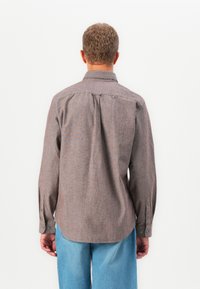 Hombre con cabello rizado corto, vistiendo una camisa marrón de manga larga y jeans de un azul claro, de pie frente a una pared blanca.