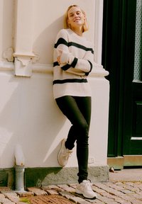 Pull crème rayé avec des rayures noires, jean skinny noir et baskets blanches. Fond texturé avec un sol en pavés.