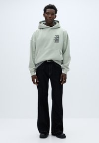 Mintgroene hoodie met een grafisch ontwerp en een voorzak. Gecombineerd met zwarte, hooggesitueerde jeans en zwarte schoenen. Simpel, ontspannen pasvorm.