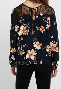 Blusa floreale navy con maniche lunghe, yoke in pizzo e dettaglio a chiave sul retro. Presenta fiori pesca e bianchi su uno sfondo scuro.