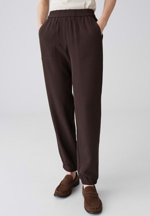 Personne portant un pantalon élastique marron foncé à la taille et aux chevilles avec poches latérales et mocassins marron sur un fond uni.