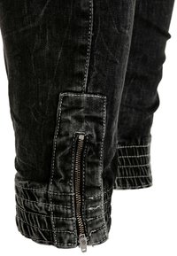 Un pantalon en denim noir présente une fermeture éclair à la cheville avec une texture côtelée et des coutures blanches le long des coutures. Le design global est ajusté.