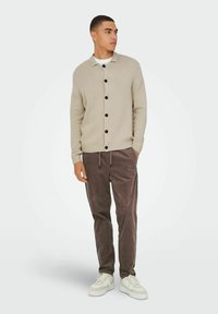 Beige strikket cardigan med sorte knapper, ribbede manchetter og kant. Sammen med brune kordfløjspants og hvide sneakers. Simpelt design, behagelig pasform.