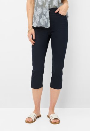 Kvinde iført marineblå cropped bukser, en mønstret ærmeløs bluse, hvide slidesandaler og et guldarmbånd på hvid baggrund.