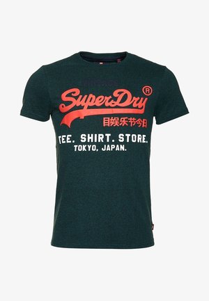 T-shirt manches courtes vert foncé avec texte rouge et blanc indiquant "SuperDry," caractères japonais, et "TEE. SHIRT. STORE. TOKYO, JAPAN."
