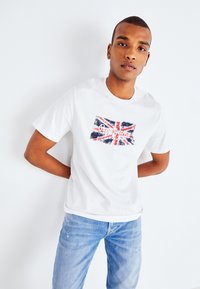Witte katoenen t-shirt met een verouderd Union Jack ontwerp in rood, blauw en wit, met in het midden van de vlag de tekst "Real Jeans".