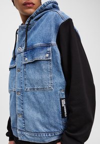 Denim jas met een blauwe voorkant en zwarte mouwen, voorzien van twee borstzakken, knoopsluitingen en een merketiket op de zijnaad.