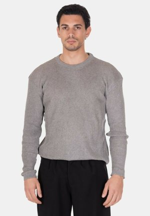 Uomo con capelli corti e scuri che indossa un maglione grigio a maniche lunghe in maglia e pantaloni neri, in piedi contro uno sfondo chiaro e uniforme.