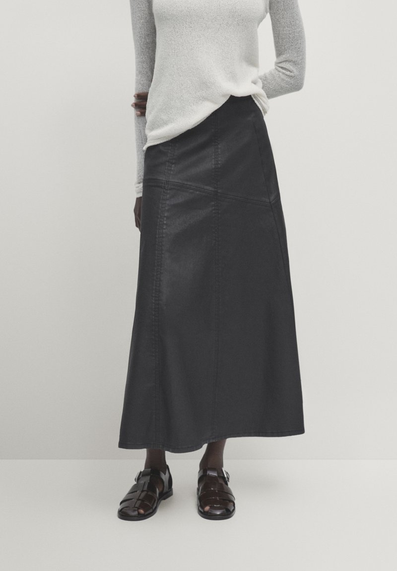 Gonna lunga in pelle nera con cuciture frontali e silhouette dritta, abbinata a sandali neri con cinturini e a un maglione grigio chiaro con texture.