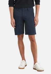 Navyblauwe katoenen shorts hebben een klassieke snit, voorzakken en een rits met knoopsluiting. Gecombineerd met witte sneakers.
