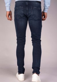 Hombre vistiendo jeans ajustados de color azul oscuro y zapatillas blancas, de pie con las manos relajadas a los lados contra un fondo neutral.