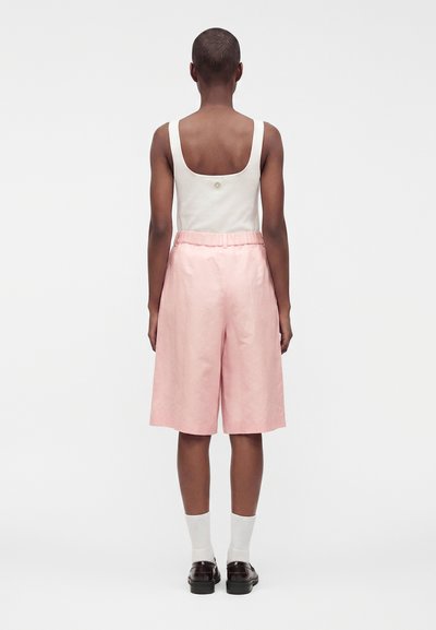 Personne aux cheveux courts se tient de dos, portant un haut blanc sans manches, un short rose clair arrivant aux genoux, des chaussettes blanches et des chaussures foncées.